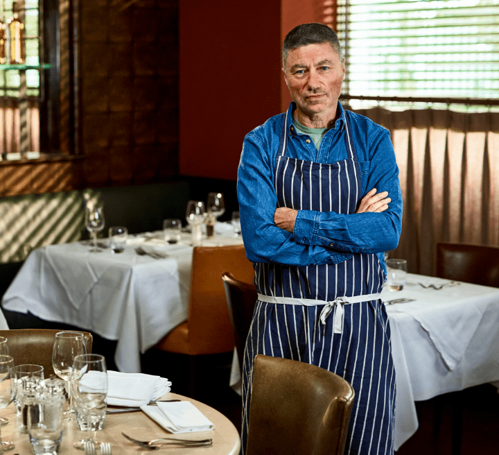 Male chef with short dark hair and moderate rosacea, standing in the empty restaurant with his arms crossed looking at the camera. Not an actual patient.