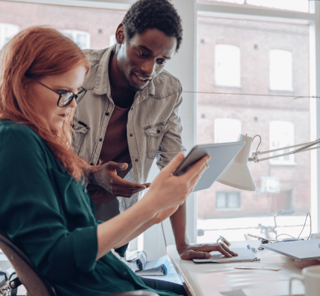 Young woman with long red hair and mild rosacea, wearing glasses looking at an iPad next to a work colleague. Not an actual patient.