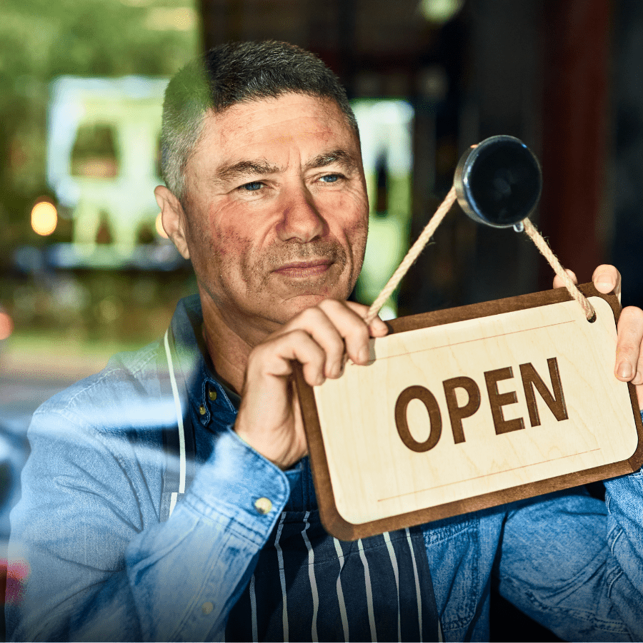 Male chef with short dark hair and moderate rosacea, looking out of the restaurant door holding the OPEN sign. Not an actual patient.