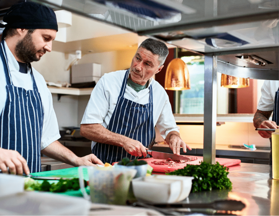 Make chef with short dark hair and moderate rosacea, talking with a colleague in the kitchen while chopping food. Not an actual patient.