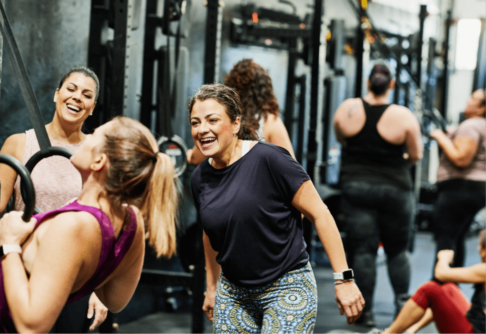 A 35 year old woman with dark curly hair and mild rosacea, is laughing with her friends at the gym