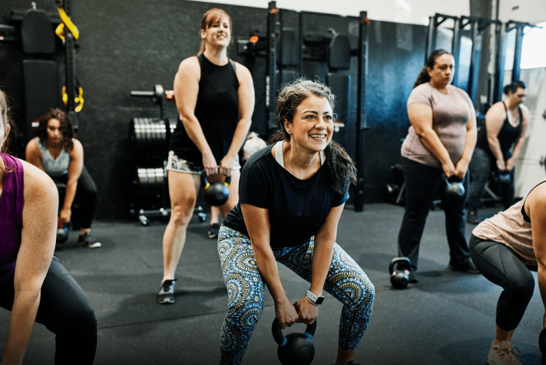 A 35 year old woman with dark curly hair and mild rosacea, is smiling while lifting a dumb bell during a class