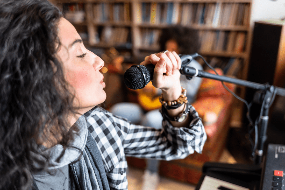 A 30 year old woman with long dark curly hair and mild rosacea, is holding a microphone to her mouth and singing