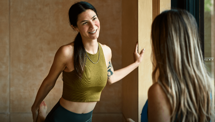 Young woman wearing yoga outfit during a class, is stretching her leg while talking to a friend and is almost clear of rosacea