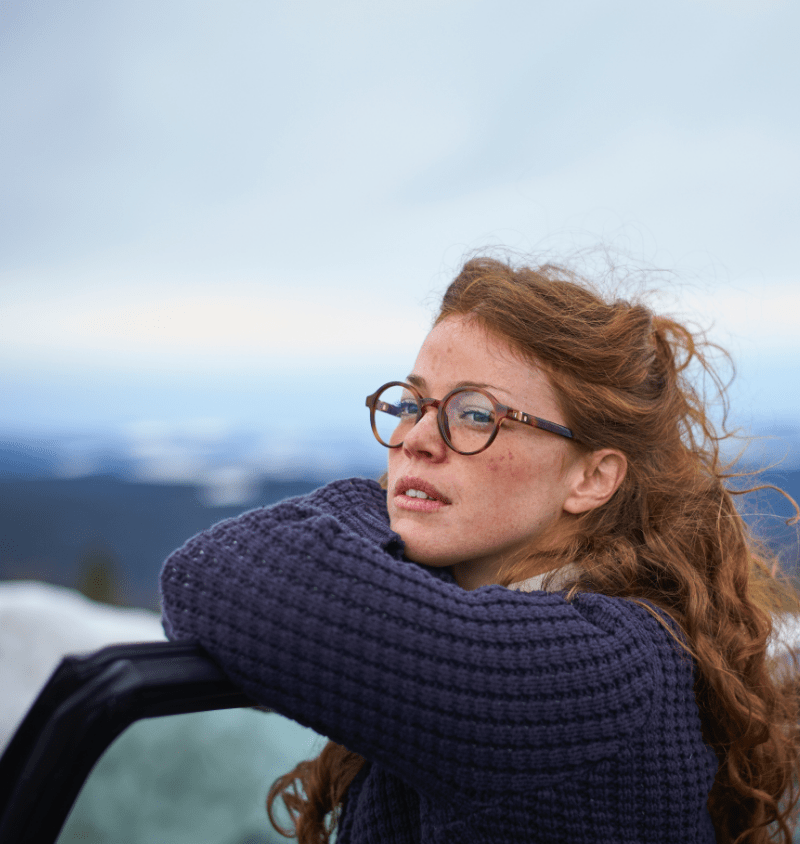 Outside image of  red haired girl wearing glasses and jumper showing mild to moderate rosacea on her cheeks ad forehead